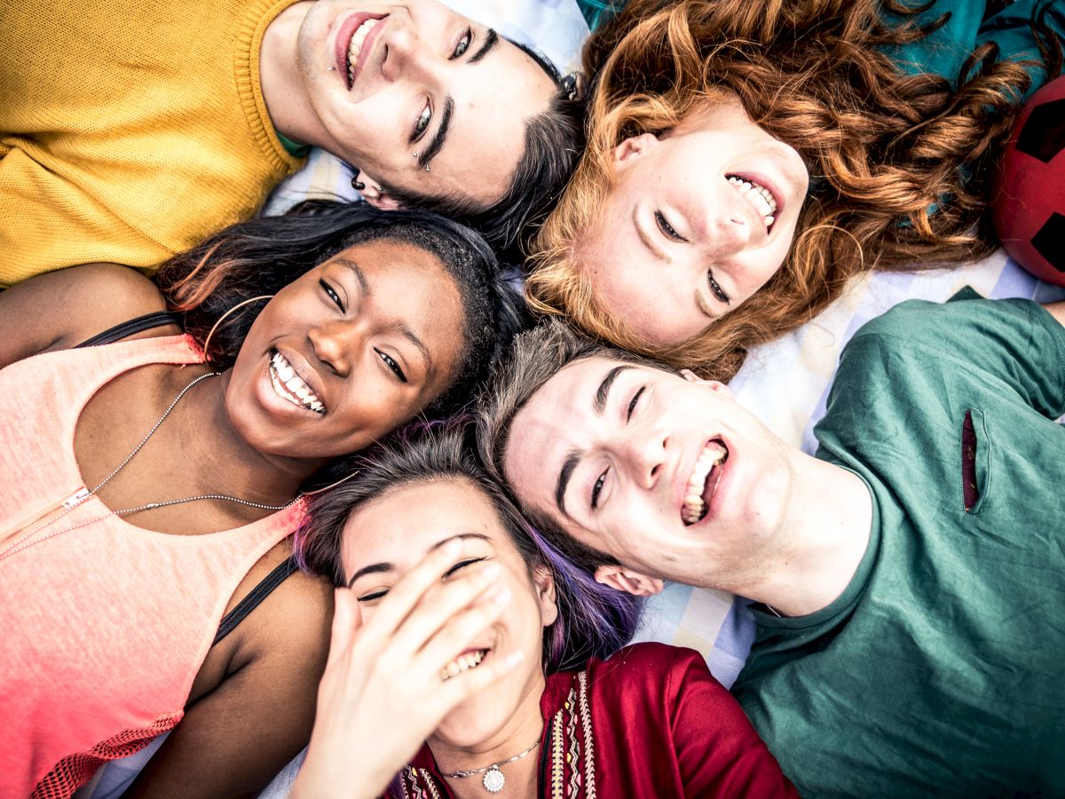A group of five people lying in a circle on grass, smiling and laughing, with a red soccer ball nearby on the right.