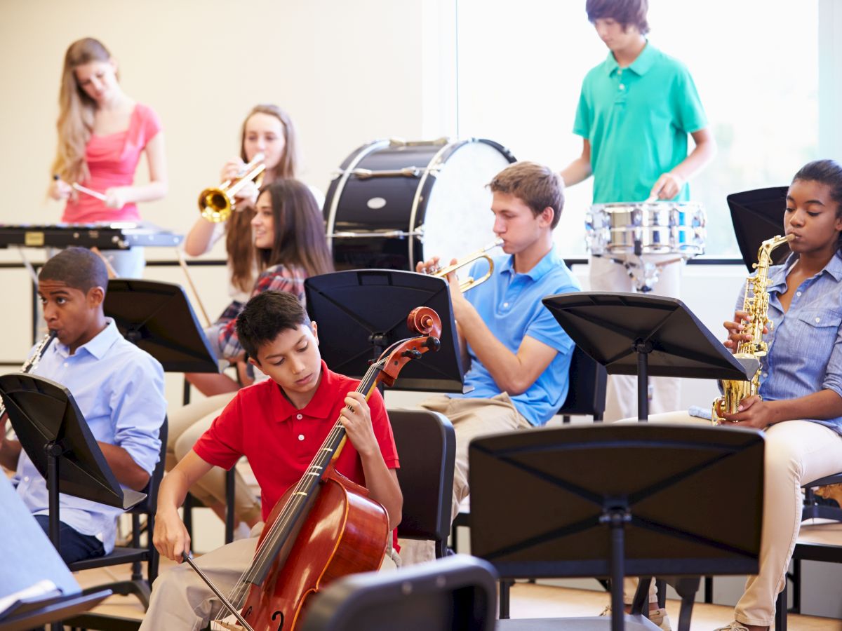 A group of students is playing various instruments in a band rehearsal, including a cello, saxophone, trumpet, and percussion.